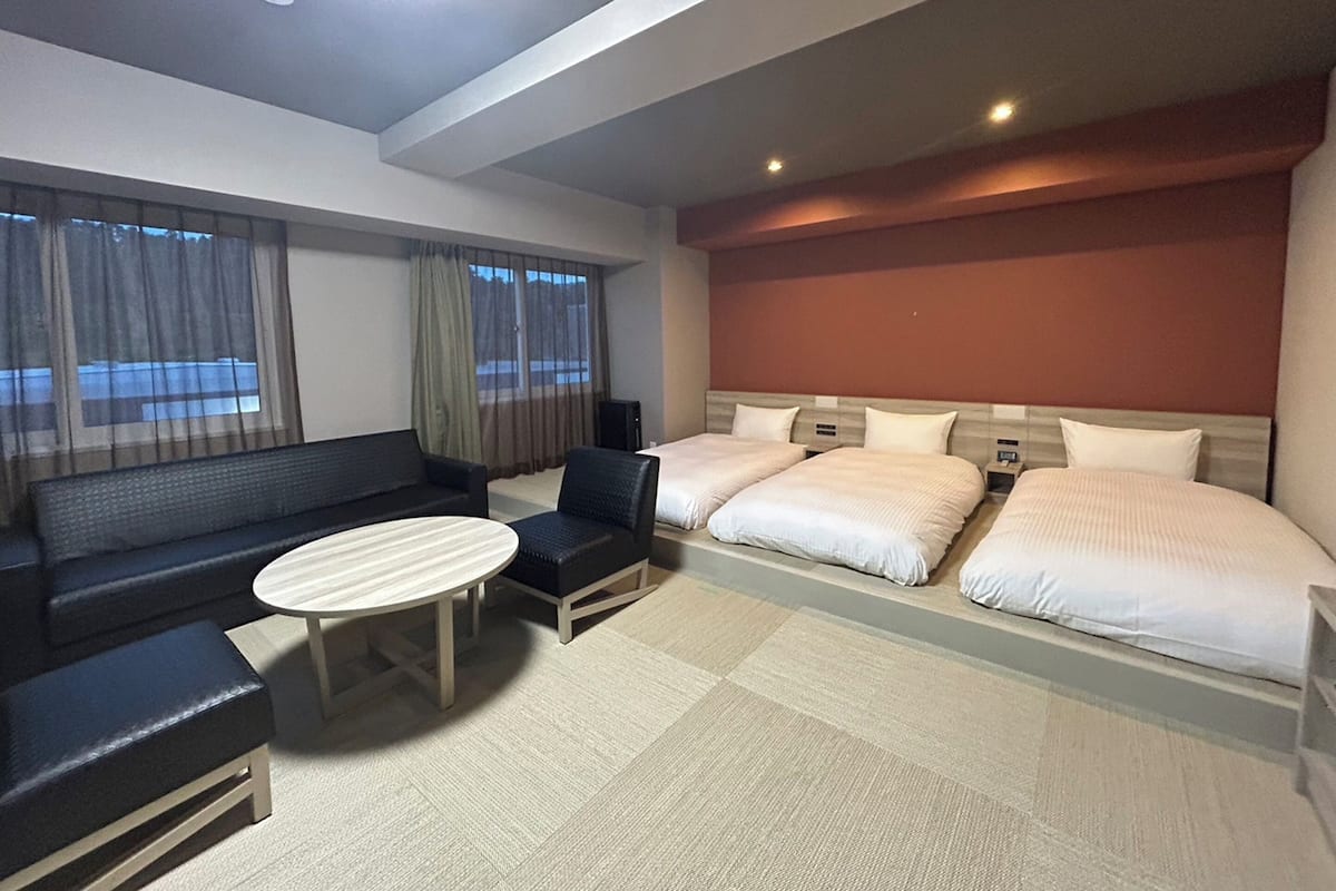 A hotel room with a sofa, two single beds, a small table, and a large window with curtains.