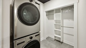 A laundry room with a front-loading washer and dryer, a white closet with shelves and hanging space, and a grey carpeted floor.