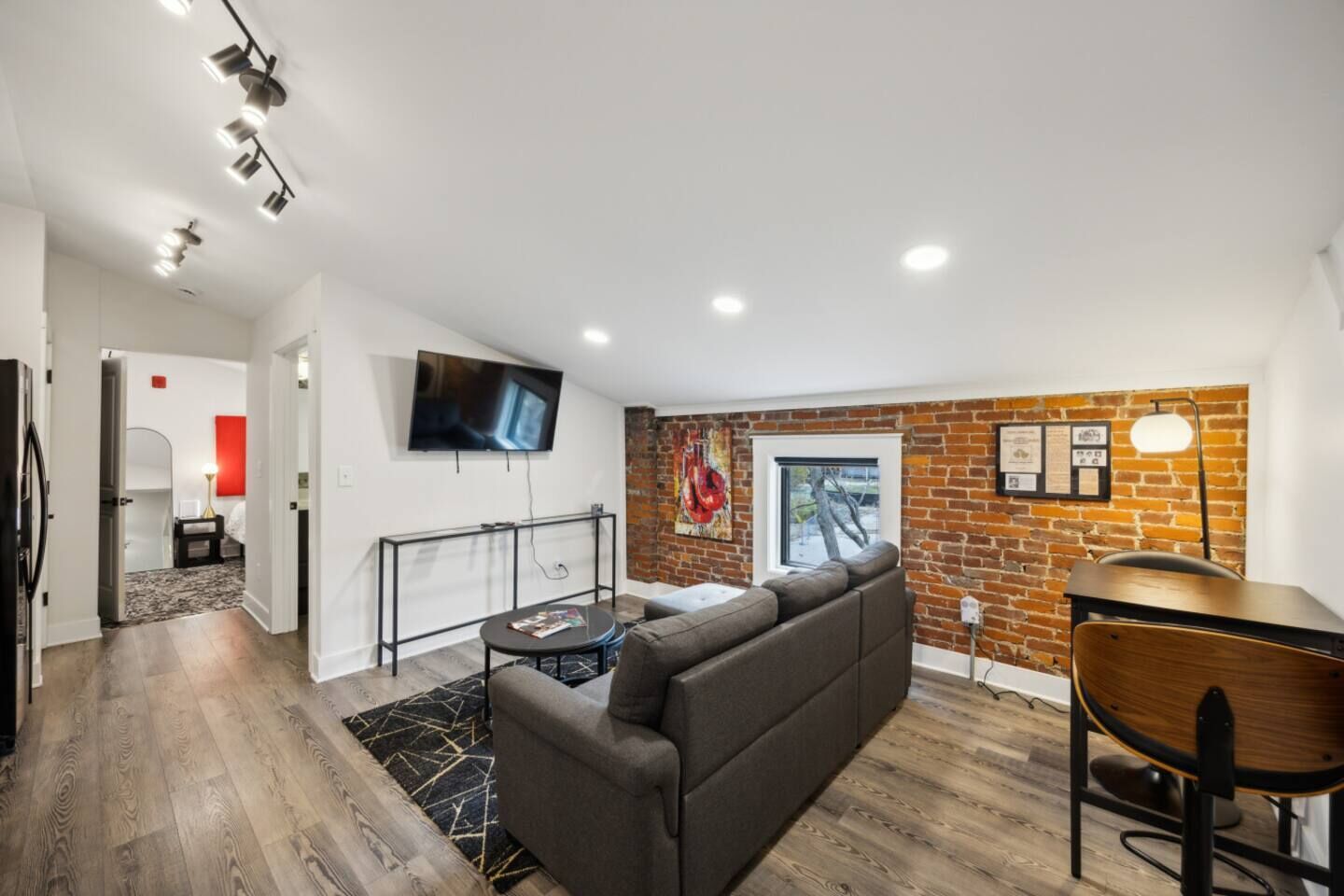 A modern living room with a sectional sofa, a TV mounted on the wall, a brick accent wall, and a dining area with a table and chairs.