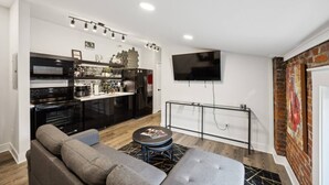 Ein modernes Wohnzimmer mit grauem Sofa, einem Couchtisch und einer Küchenzeile mit schwarzen Geräten sowie einem an der Wand befestigten Fernseher.