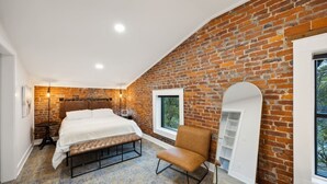 Un dormitorio con pared de ladrillo, una cama, un banco, una silla y una ventana.