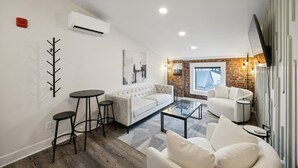 Una sala de estar moderna con un sofá blanco, una mesa de centro redonda y un sillón blanco. Se aprecia una pared de ladrillo y una ventana al fondo.