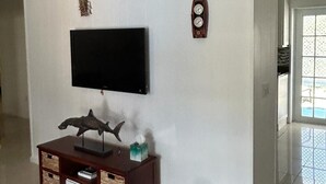 A living room with a flat-screen TV mounted on the wall, a wooden entertainment unit below it, a decorative wall clock, and a patterned vase on the floor. - Endless Summer, Cozy Bahamian Cottage (Man o War Cay)