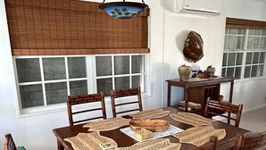 A dining area with a wooden table and chairs, a hanging light fixture, and a window with blinds. - Endless Summer, Cozy Bahamian Cottage (Man o War Cay)