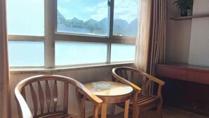 Una habitación con dos sillas de madera y una pequeña mesa redonda, una ventana con vista a montañas y un lago, y una planta en un estante.