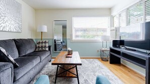 Una sala de estar moderna con un sofá gris, una mesa de centro de madera, un mueble para TV y un ventanal con persianas.
