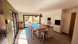 Een moderne keuken met een centraal eiland, barstoelen en uitzicht op een zwembad door grote schuifdeuren.