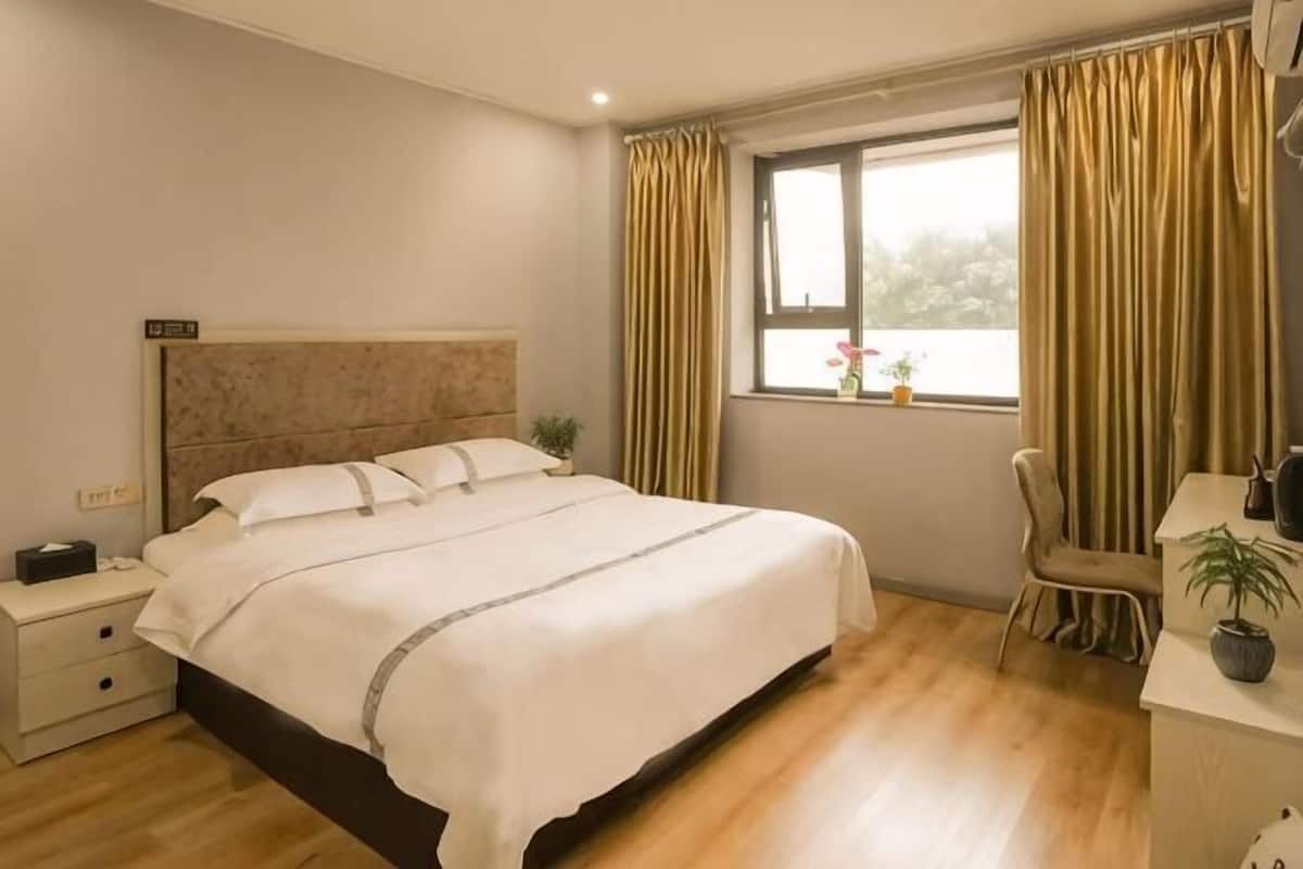 A bedroom with a bed, bedside table, chair, window, curtains, and a small table with a plant.