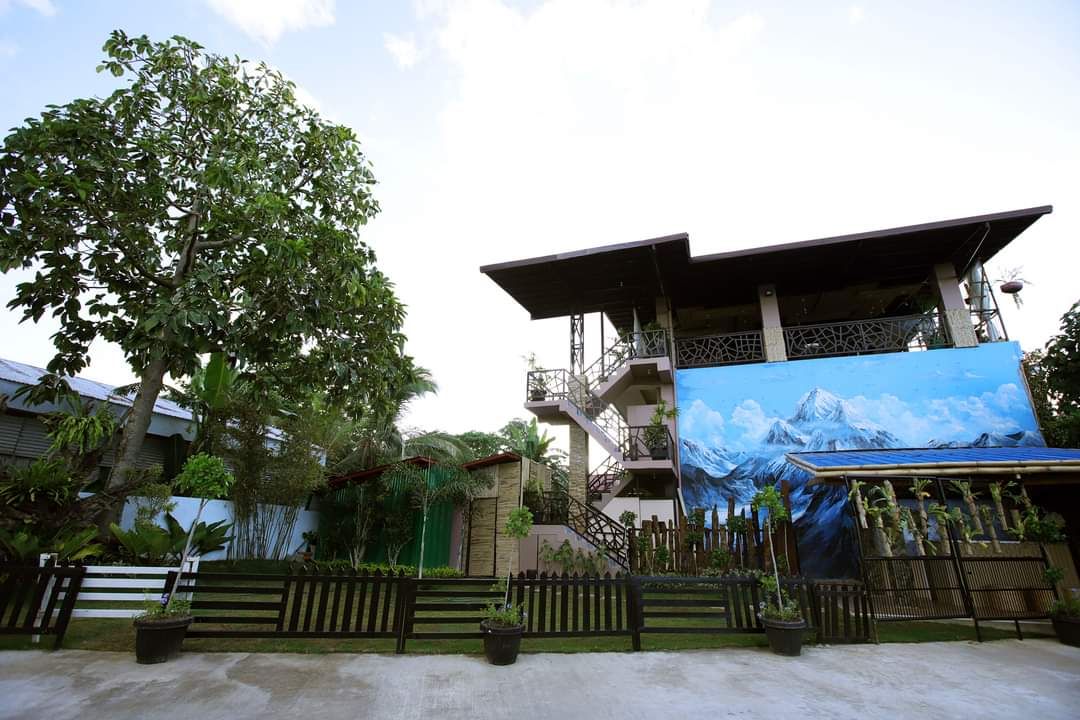 A building with a large mural of a mountain landscape, surrounded by lush greenery and a wooden fence.