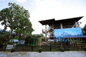 Un edificio con un gran mural de un paisaje montañoso, rodeado de exuberante vegetación y una valla de madera.