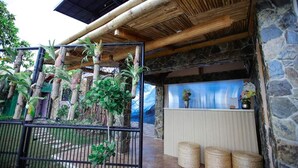 Un espace extérieur rustique agrémenté d’un mur en pierre, d’un plafond en bois et d’une clôture en bambou. On y trouve des paniers en osier et un petit coin salon.