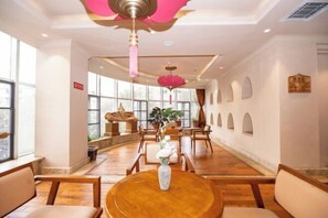 A hotel lobby with wooden flooring, round tables, and chairs. There are large windows allowing natural light, and a ceiling fan with a pink decorative element. - Liaokuo Jingshe Hotel (Qujing)