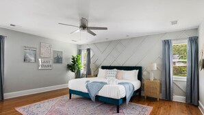Un dormitorio con cama, ventilador de techo, mesita de noche, ventana con cortinas y una pared decorada con cuadros.