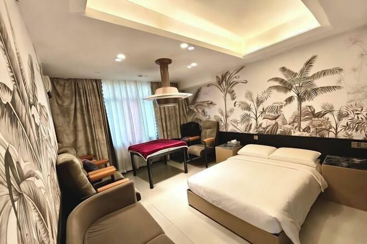 Habitació d'hotel moderna amb un llit gran, una zona de seients amb una cadira i una taula petita, una làmpada de sostre i un mural a la paret.