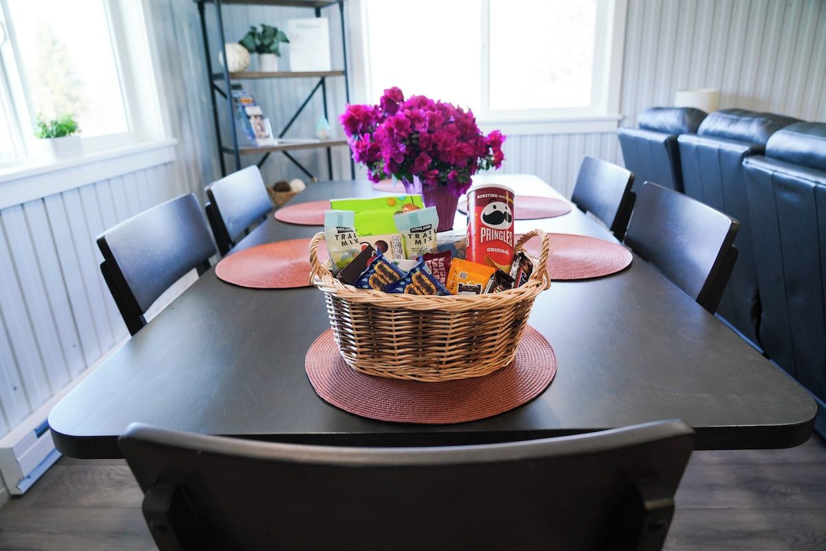 A dining table with a basket of snacks, a vase of flowers, and a bookshelf in the background.