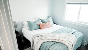 A neatly made bed with a white comforter and multiple pillows, a bedside lamp, and a window with blinds.