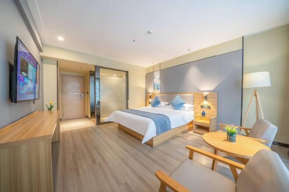 A hotel room with a bed, bedside table, lamp, chair, and a small table with a plant.