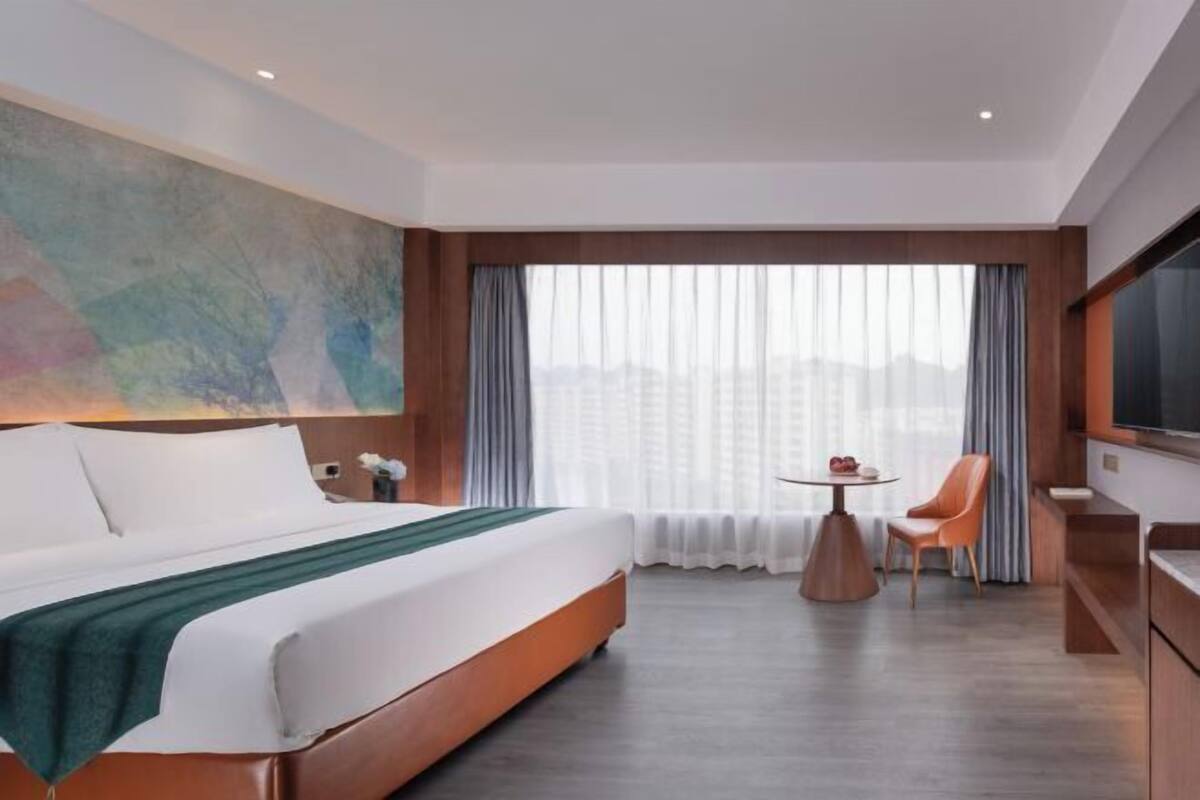 A hotel room with a bed, a table with chairs, a TV, and a large window with curtains.