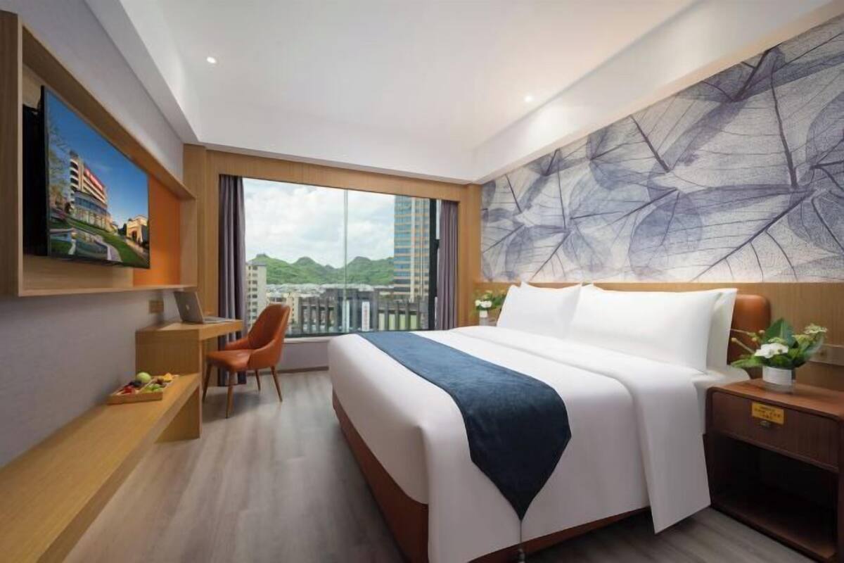 A hotel room with a large bed, a desk with a laptop, a chair, a TV mounted on the wall, and a view of the cityscape.