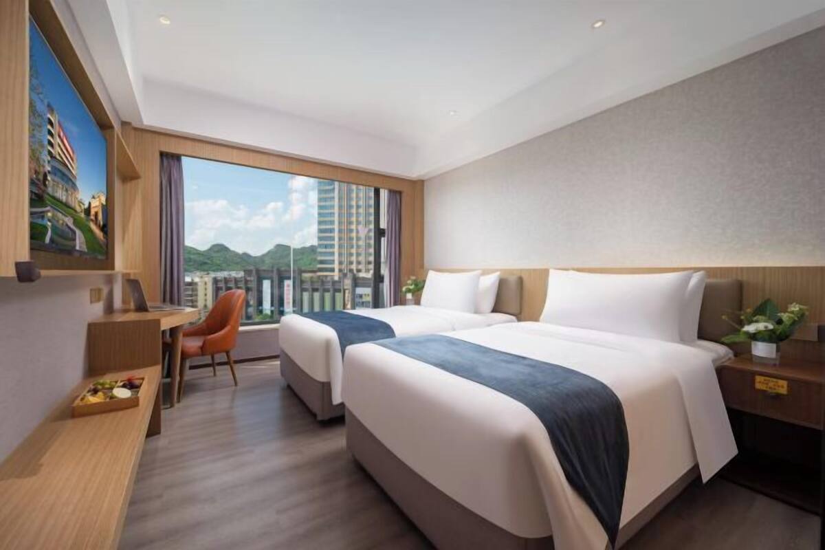 A hotel room with two beds, a desk, a chair, a TV, and a large window with a city view.