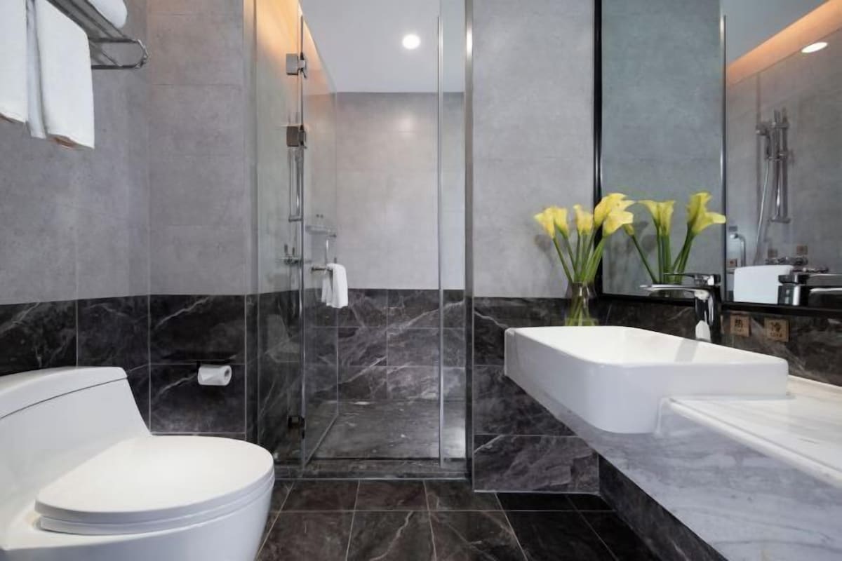Un baño moderno con ducha envuelta en vidrio, inodoro blanco, una bañera blanca grande y un espejo con un jarrón de flores amarillas.