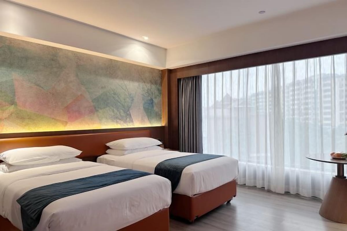 Habitación de hotel con dos camas, un cuadro abstracto grande en la pared, un ventanal con cortinas y una mesa redonda con un florero.