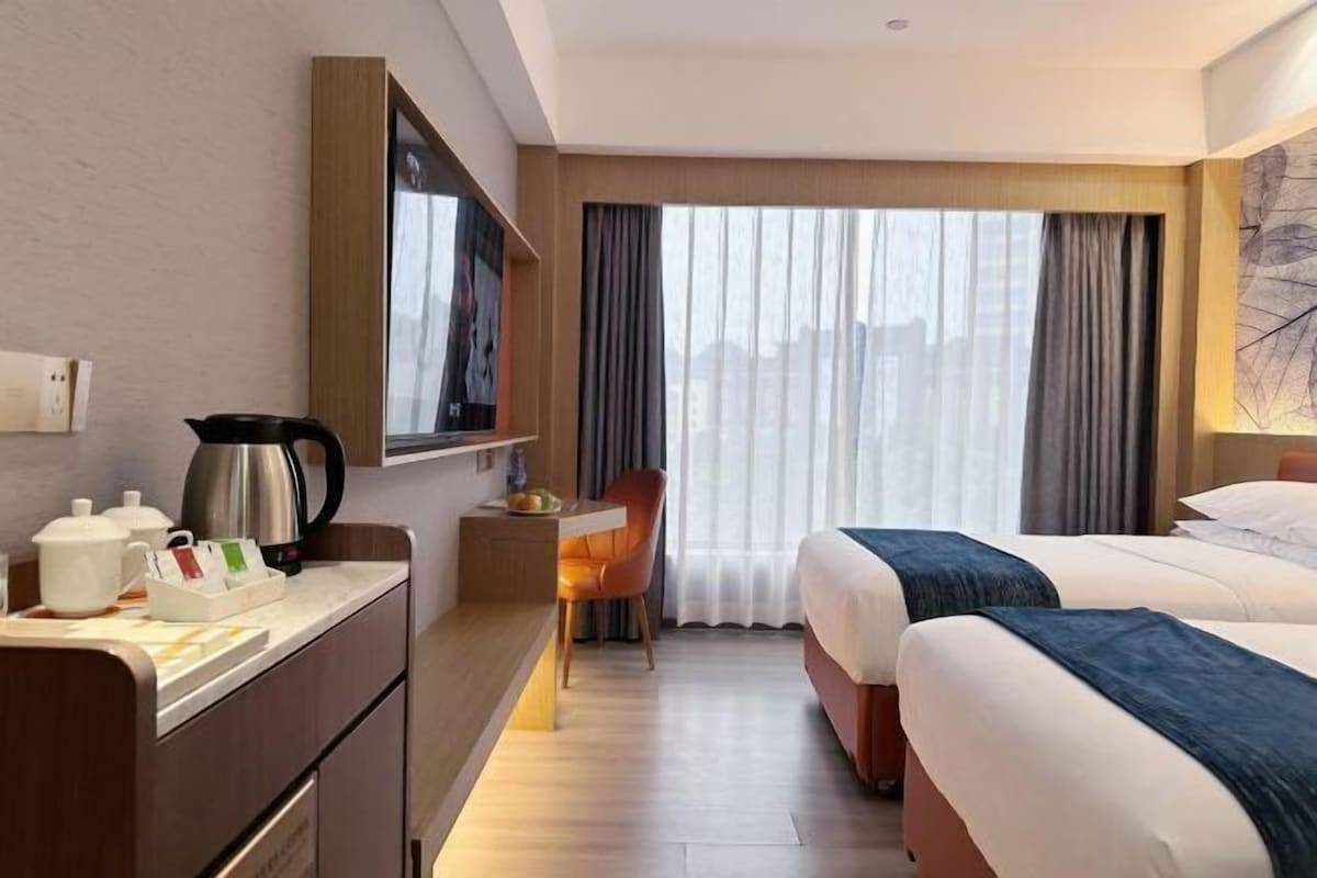 Una habitación de hotel con cama, escritorio con hervidor, una silla y ventana con cortinas.