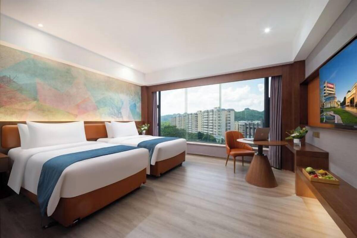 A hotel room with two beds, a desk, a chair, a large window with a city view, and a painting on the wall.