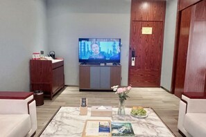 Une chambre d’hôtel avec un téléviseur posé sur un meuble en bois, une table basse en marbre ornée d’un vase de fleurs, une télécommande et une petite table sur laquelle se trouvent une boîte de mouchoirs et une assiette de fruits.
