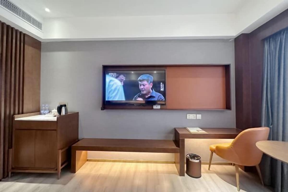 Habitación de hotel con televisión montada en la pared, un banco de madera, una mesita, una silla y un cesto de basura.
