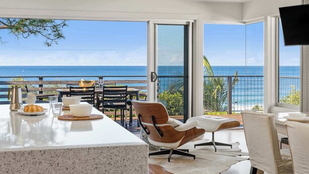 A modern kitchen with a large island, a dining area with chairs and tables, and a view of the ocean. - Surf Watch Mollymook (Mollymook Beach)