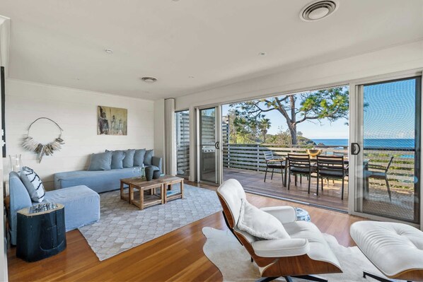 A modern living room with a sofa, coffee table, and chairs, a dining area with a table and chairs, and a large sliding glass door leading to a deck with a view of the ocean. - Surf Watch Mollymook (Mollymook Beach)