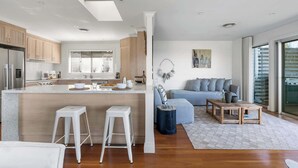 A modern kitchen with a central island, wooden cabinets, and a dining area with a sofa and coffee table. - Surf Watch Mollymook (Mollymook Beach)