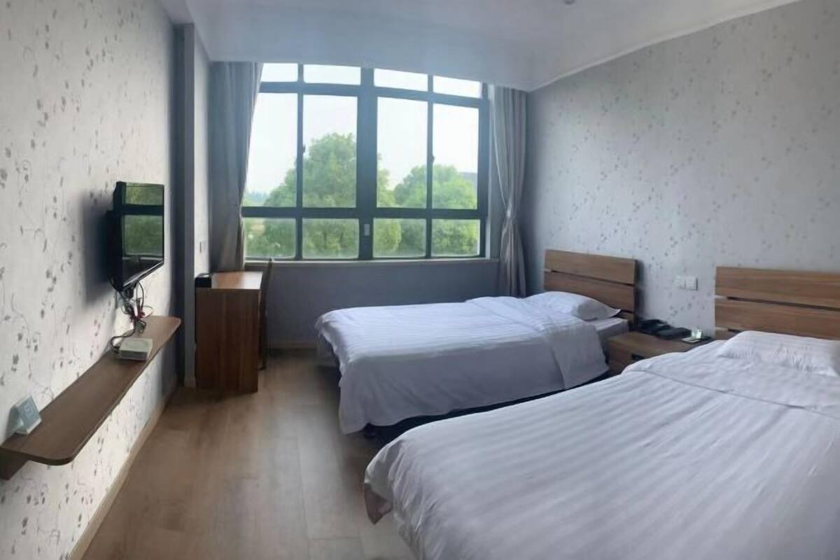 Ein Hotelzimmer mit zwei Betten, einem Fernseher auf einem Regal, einem Fenster mit Vorhängen und einem hölzernen Kopfteil.