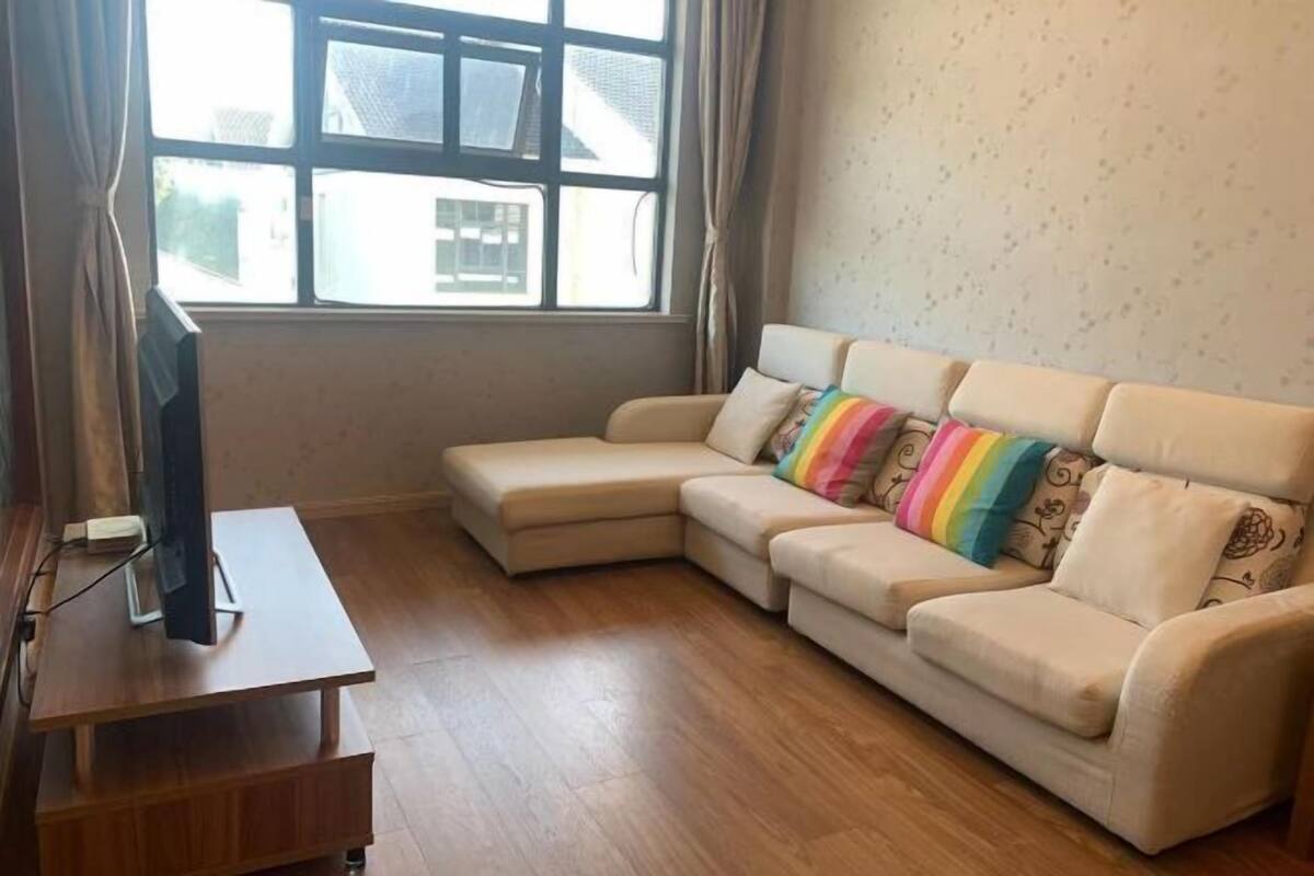 Ein Wohnzimmer mit einem beigefarbenen Ecksofa, einem Holz-TV-Möbel und einem Fenster mit Vorhängen.