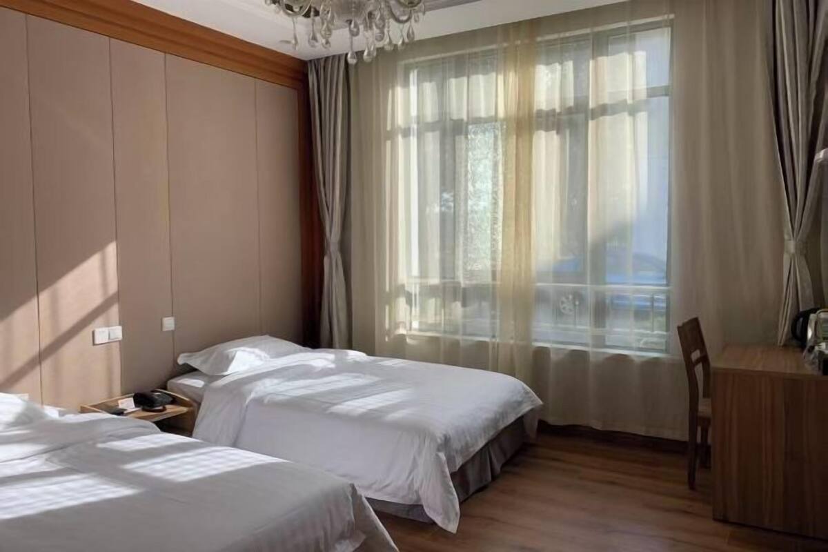 Ein Hotelzimmer mit zwei Betten, einem Schreibtisch und einem großen Fenster mit durchsichtigen Vorhängen.