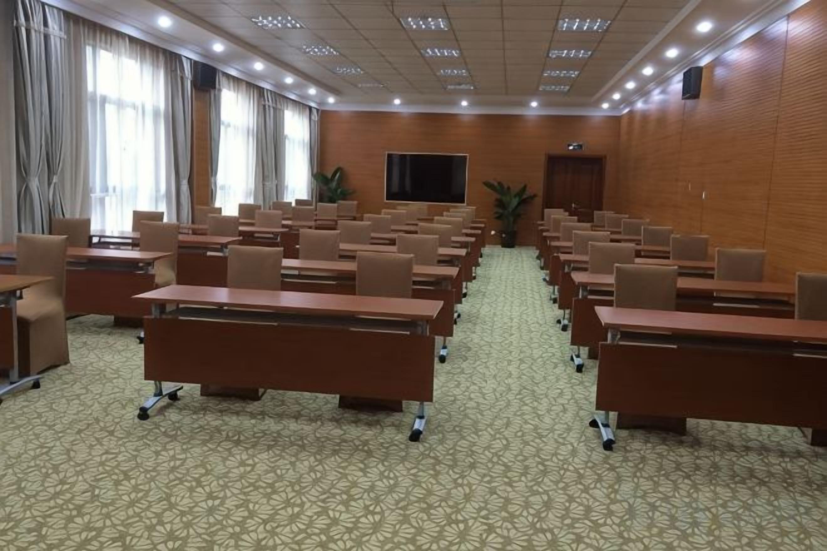 A conference room with rows of tables and chairs, a large screen, and a door in the background.