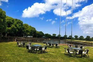 Un área con mesas y sillas al aire libre, rodeada de un campo de césped y árboles.