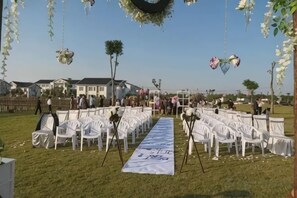 Una ceremonia de boda al aire libre con sillas blancas dispuestas en filas, un pasillo blanco y decoraciones colgantes.