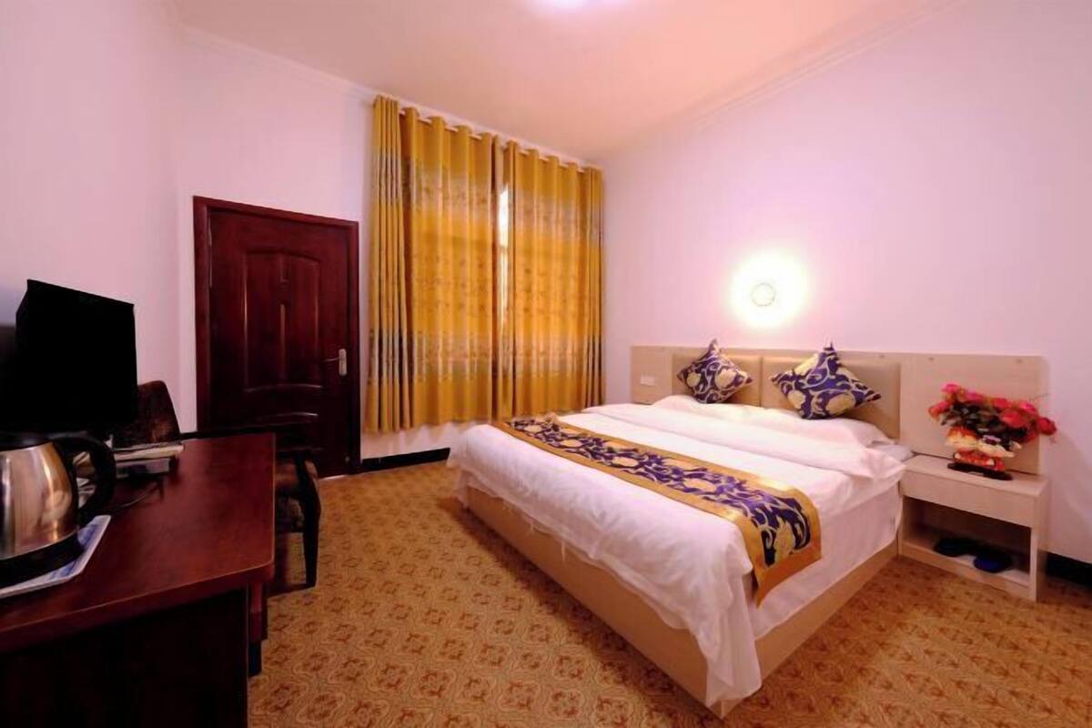 A hotel room with a bed, a desk with a kettle, a chair, a wardrobe, a nightstand with a lamp, and a window with curtains.