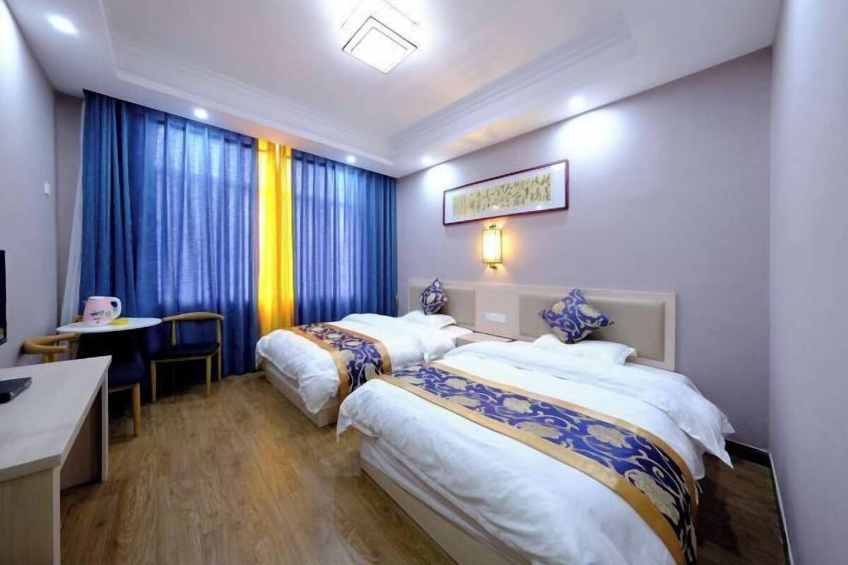 A hotel room with two beds, a desk, a chair, a table with a pink mug, and a wall-mounted lamp.