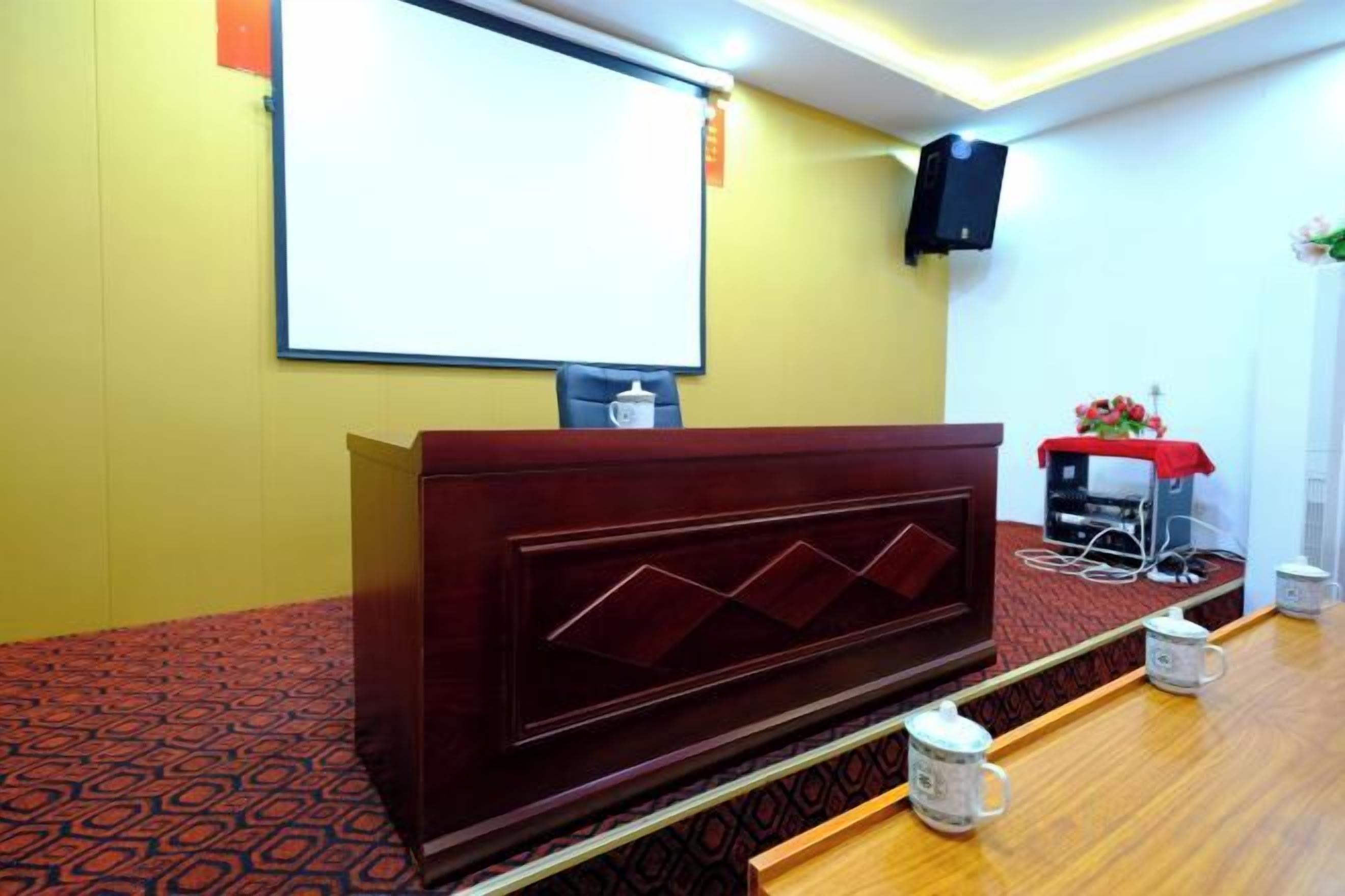 Un salón de conferencias con un atril de madera, una pantalla de proyector y un altavoz en la pared.