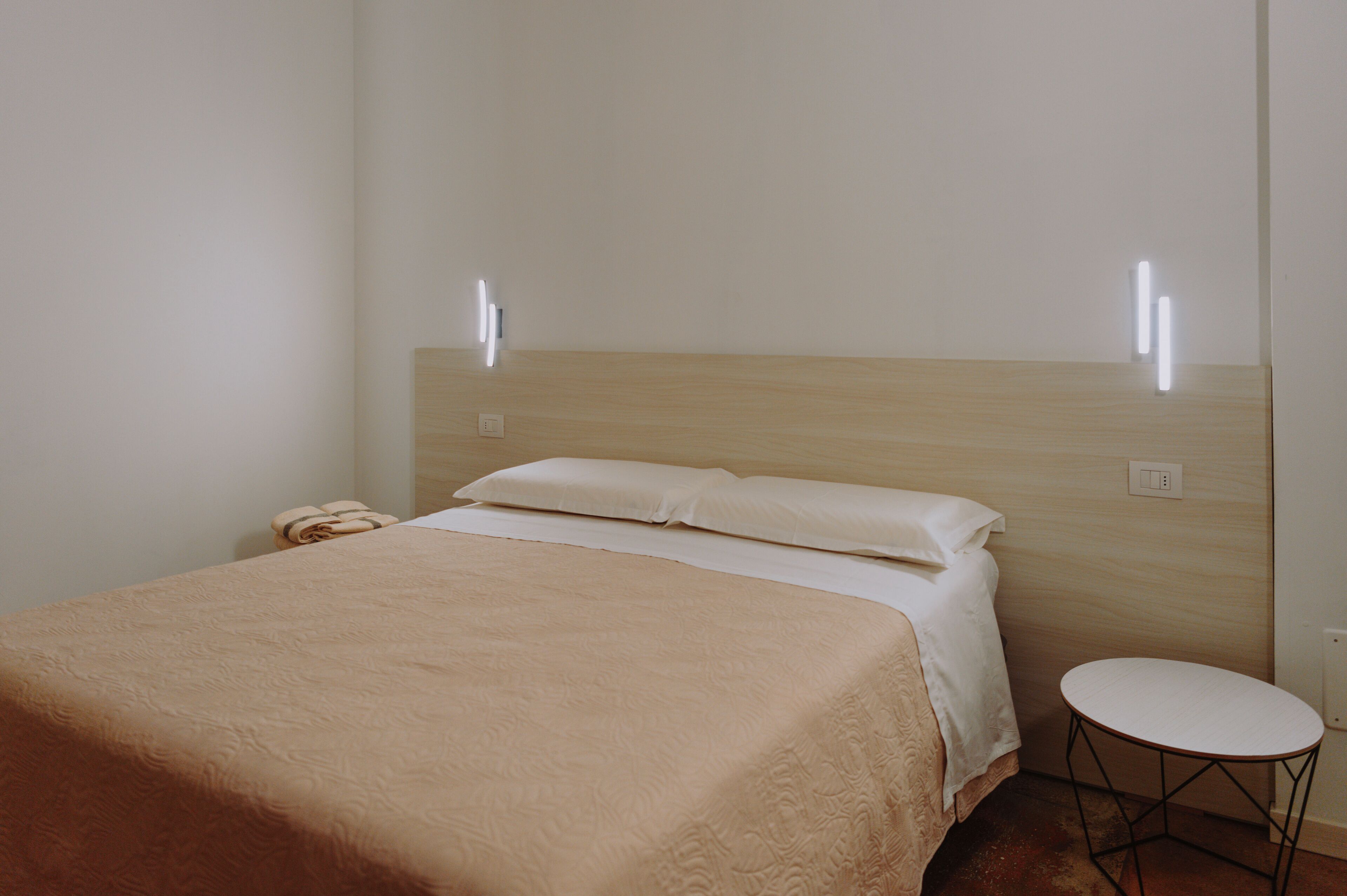 A neatly made bed with a beige blanket, a small bedside table, and a wall-mounted light fixture.