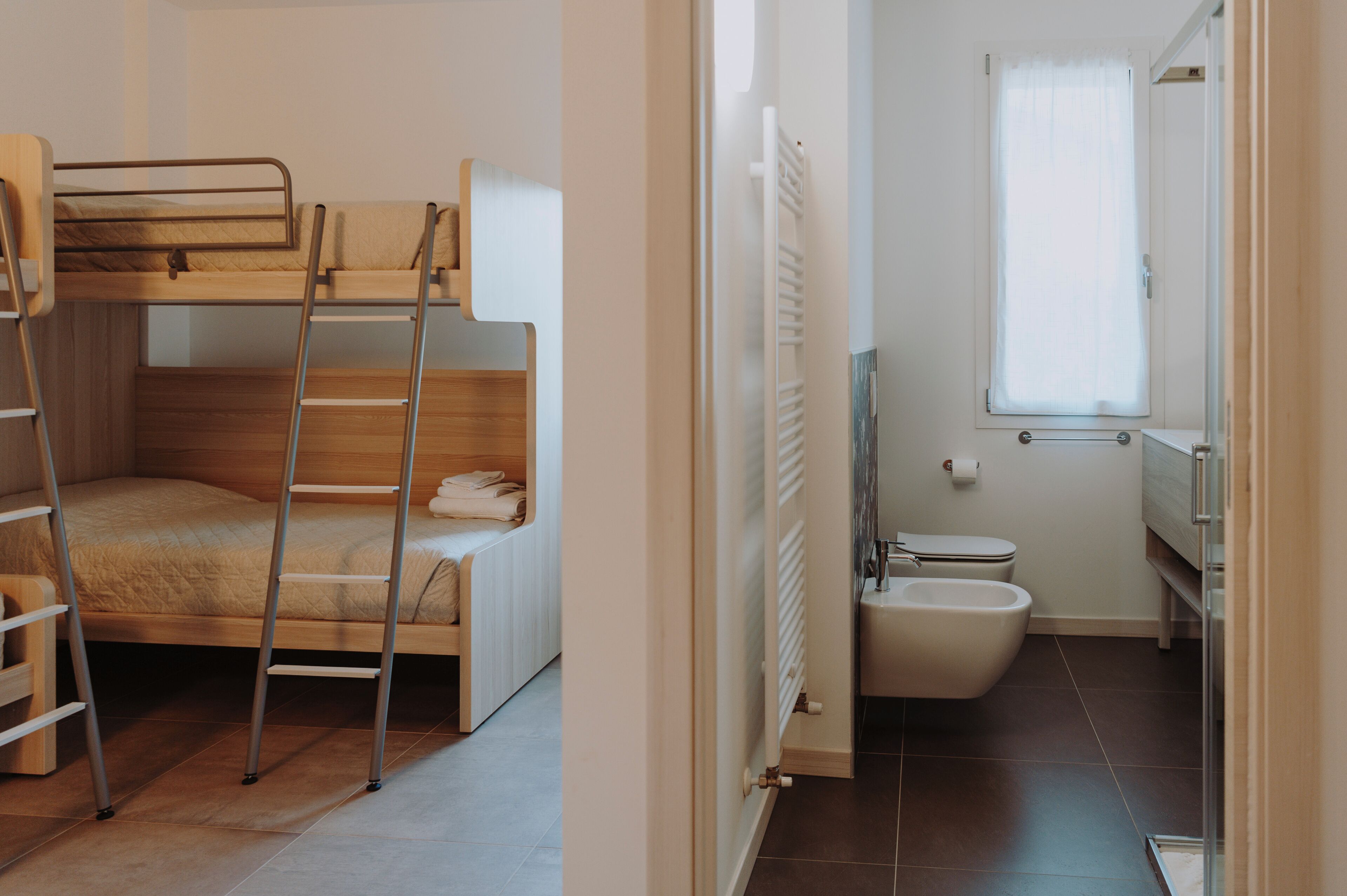 Un lit superposé avec une échelle, une petite chaise en bois et un radiateur dans une salle de bain équipée de toilettes et d’un bidet.