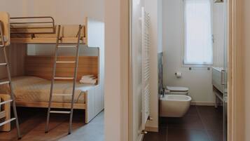 Un lit superposé avec une échelle, une petite chaise en bois et un radiateur dans une salle de bain équipée de toilettes et d’un bidet.