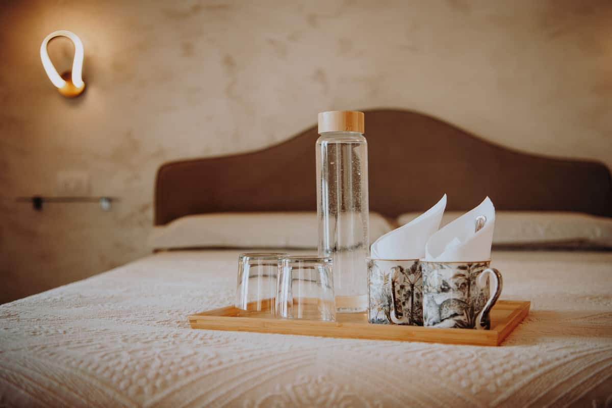 Ein ordentlich angerichtetes Frühstücksbrett auf einem Bett mit einer klaren Glasflasche, zwei Gläsern und einer dekorativen Tasse.