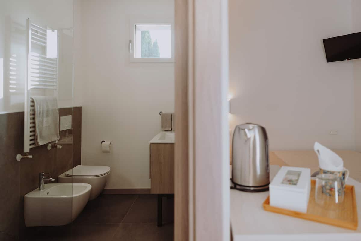 Ein modernes Badezimmer mit integriertem Bidet, Toilette und Waschbecken. Über der Toilette befindet sich ein Fenster, ein Handtuchhalter und ein kleiner Tisch mit Wasserkocher, einer Schachtel und einem Glas.