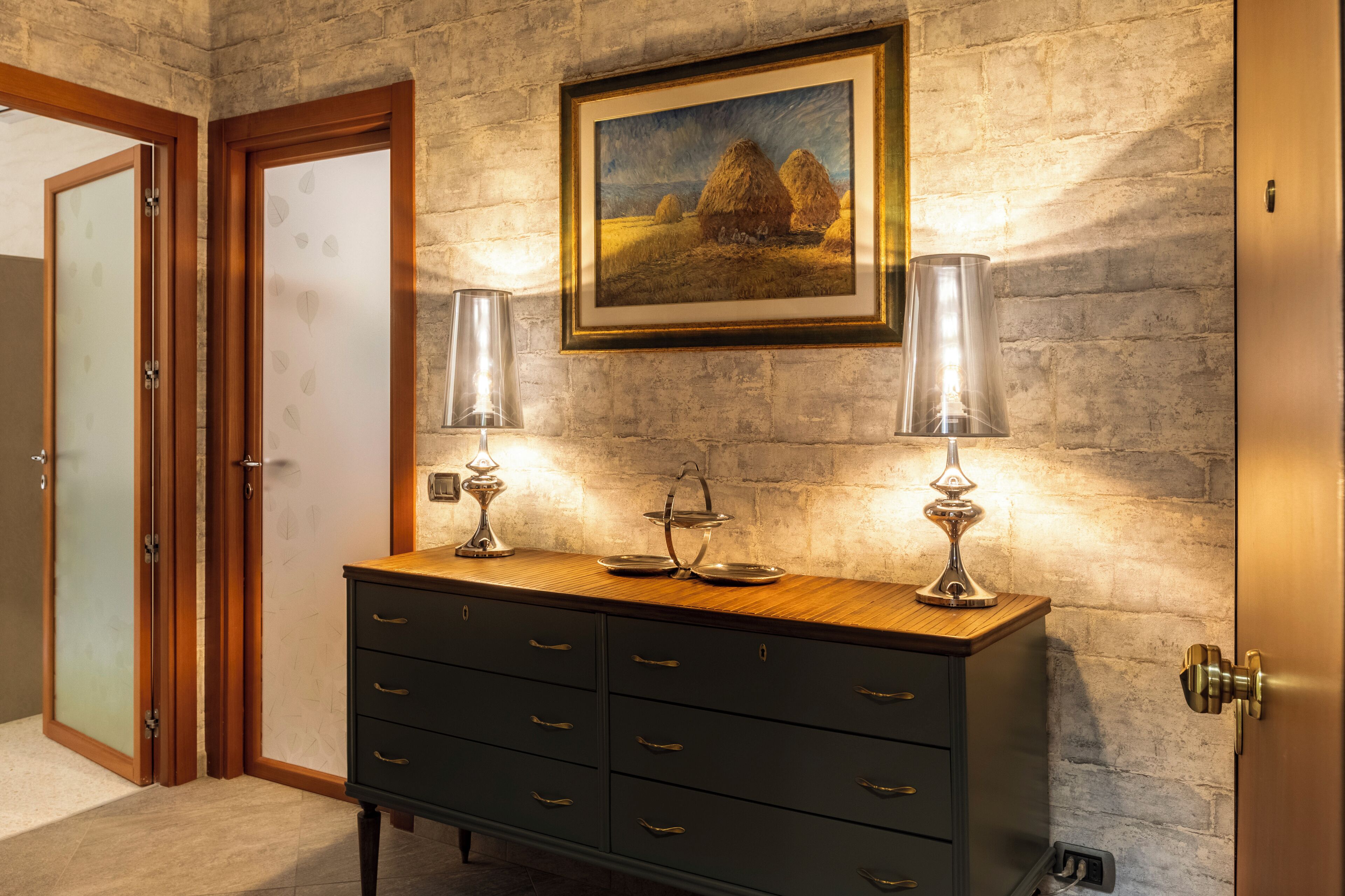 A room with a wooden cabinet, two wall-mounted lamps, and a framed painting of haystacks.
