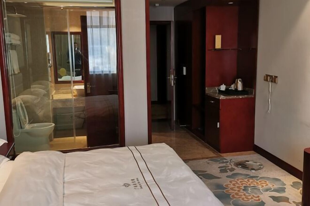 A hotel room with a bed, a bathroom with a glass shower enclosure, a small kitchenette, and a door leading to another room.