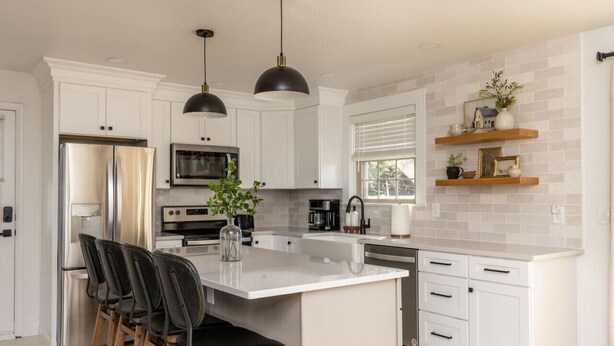 Una cocina moderna con armarios blancos, electrodomésticos de acero inoxidable, una isla grande con taburetes altos, iluminación colgante y estantes decorativos.